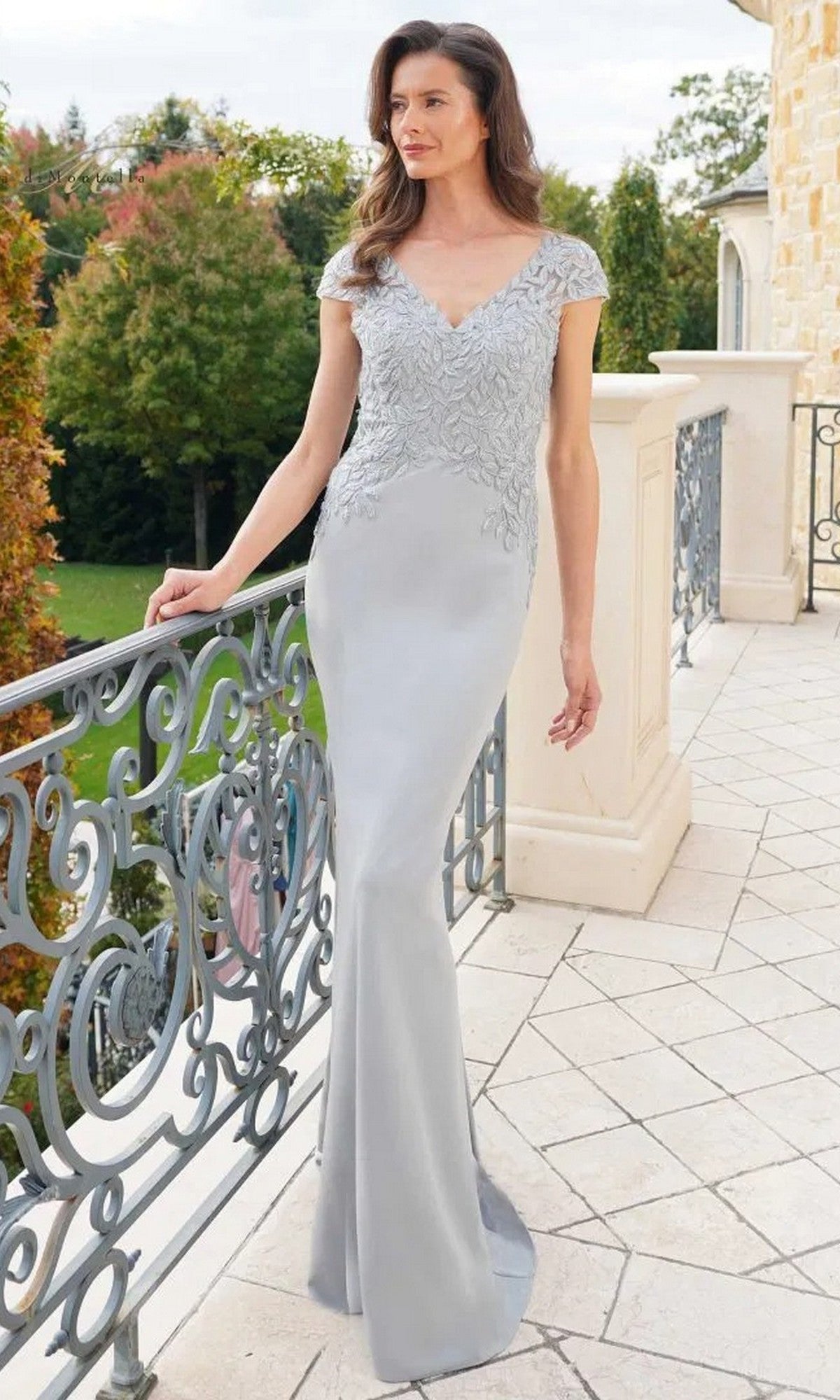 A woman stands on a balcony wearing the Long Formal Dress: Rina Di Montella RD2718 in Light Silver, featuring a v-neckline and embroidered bodice, with stone flooring, decorative railing, and trees in the background.